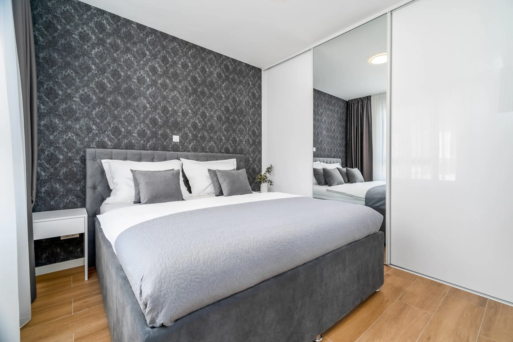 A bedroom with a gray upholstered bed, patterned accent wall, mirrored wardrobe, and wood flooring.
