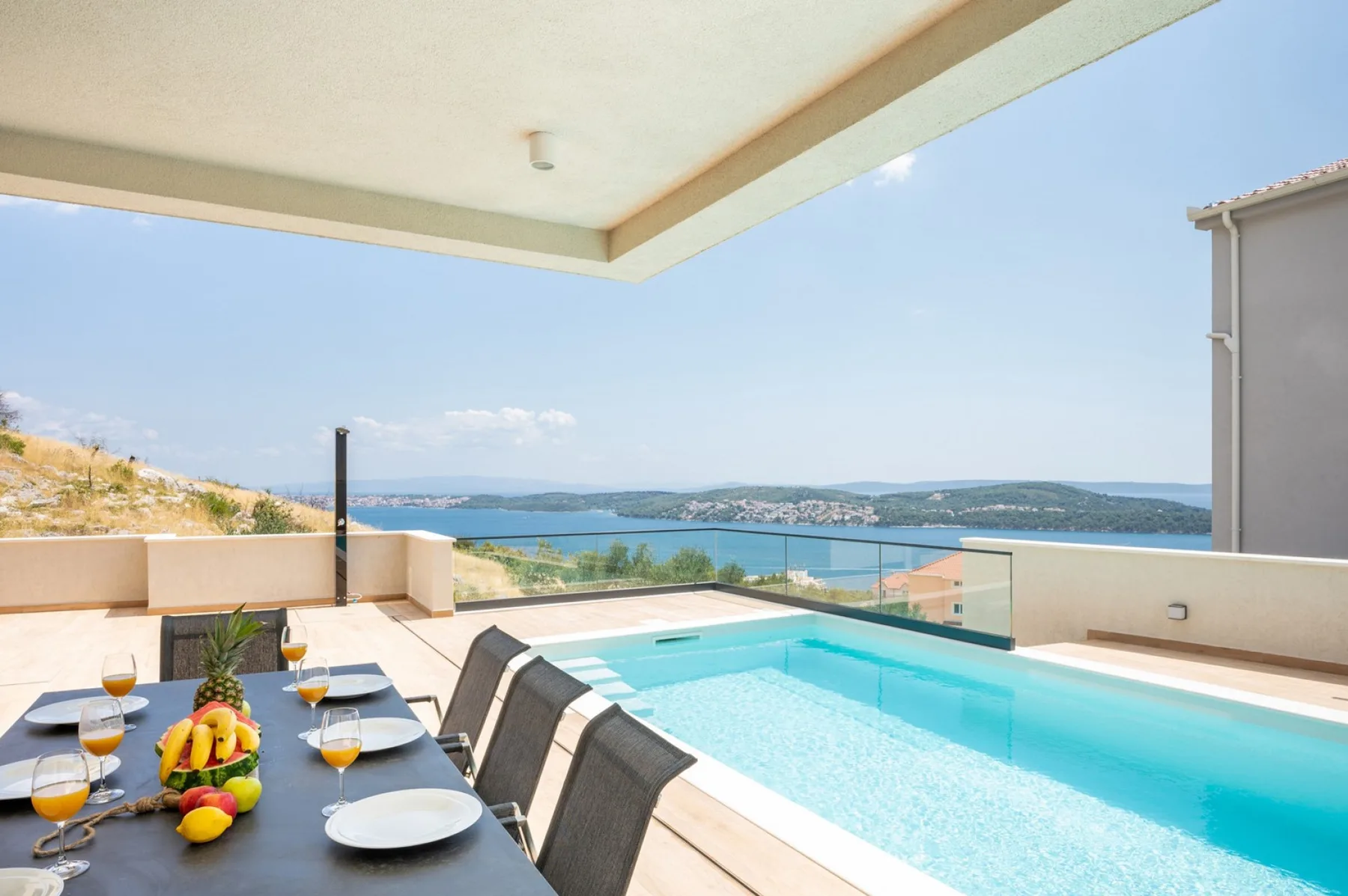 A rooftop terrace has a long turquoise pool, a set dining table, and a wide view of the sea.