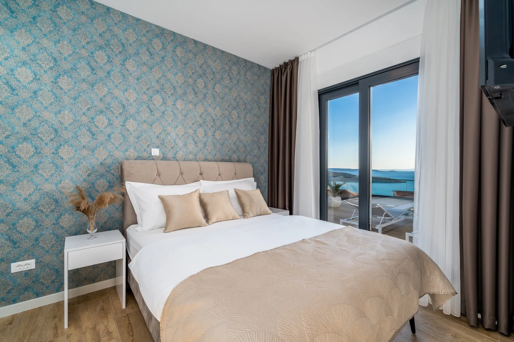 A bedroom with a beige upholstered bed, patterned blue wall, and glass doors opening onto a balcony.