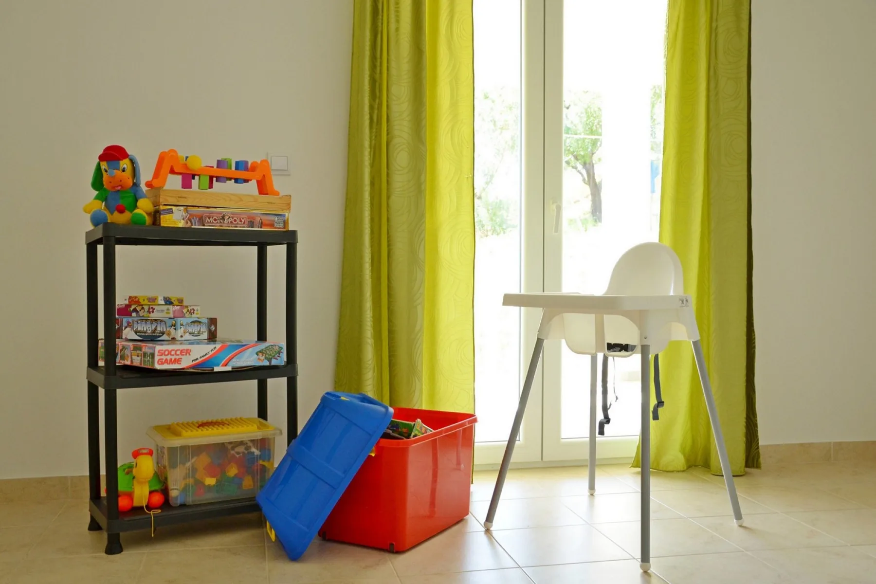 Una zona de juegos luminosa con un estante de juguetes, cajas de colores y una trona junto a la ventana.