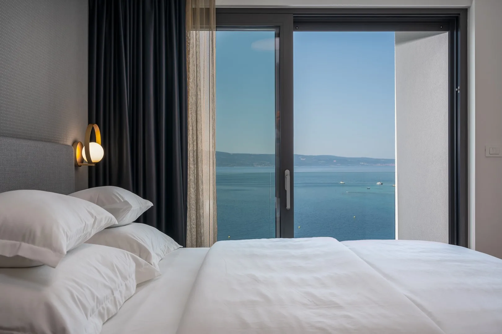 A bedroom with white bedding beside a large window overlooking the sea.
