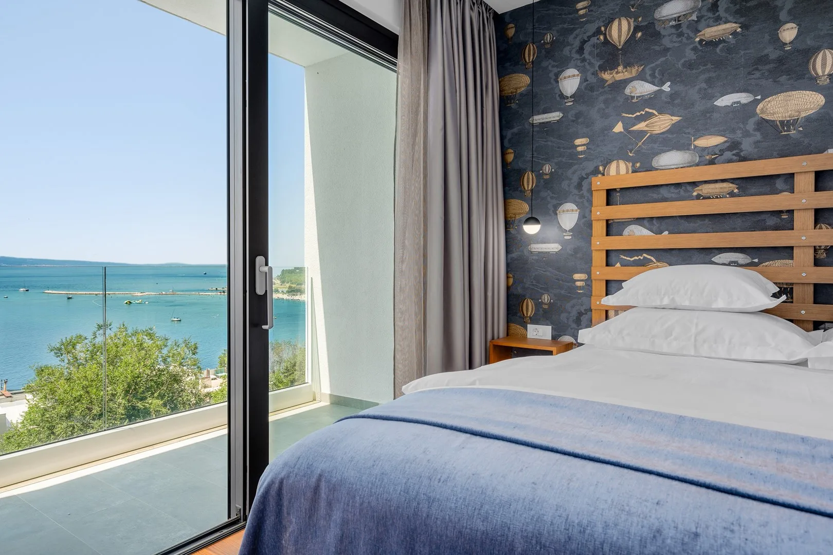 A bedroom with a bed beside a large window overlooking the sea.