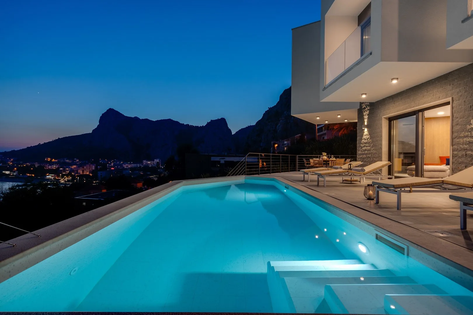 An illuminated swimming pool beside a modern building at dusk, with mountains in the background.
