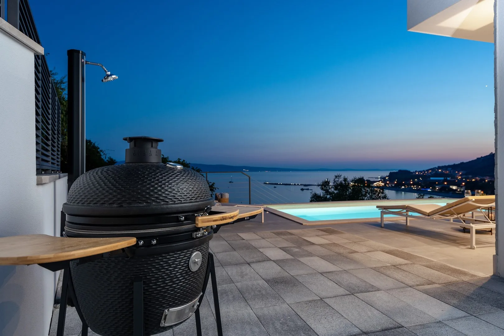 A tiled outdoor terrace overlooks a pool and coastal bay at dusk.