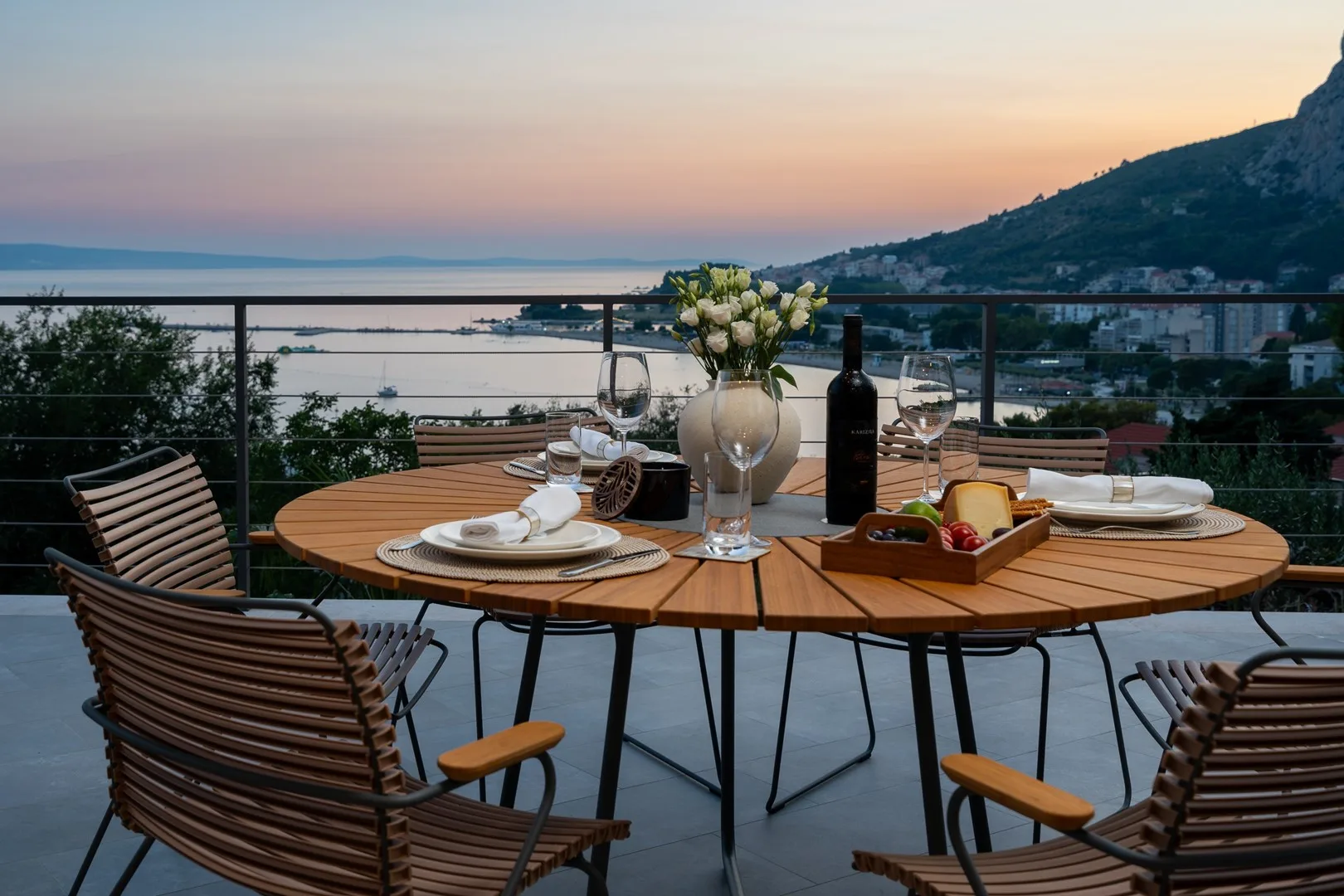 An outdoor dining table is set with plates and flowers overlooking the sea at sunset.