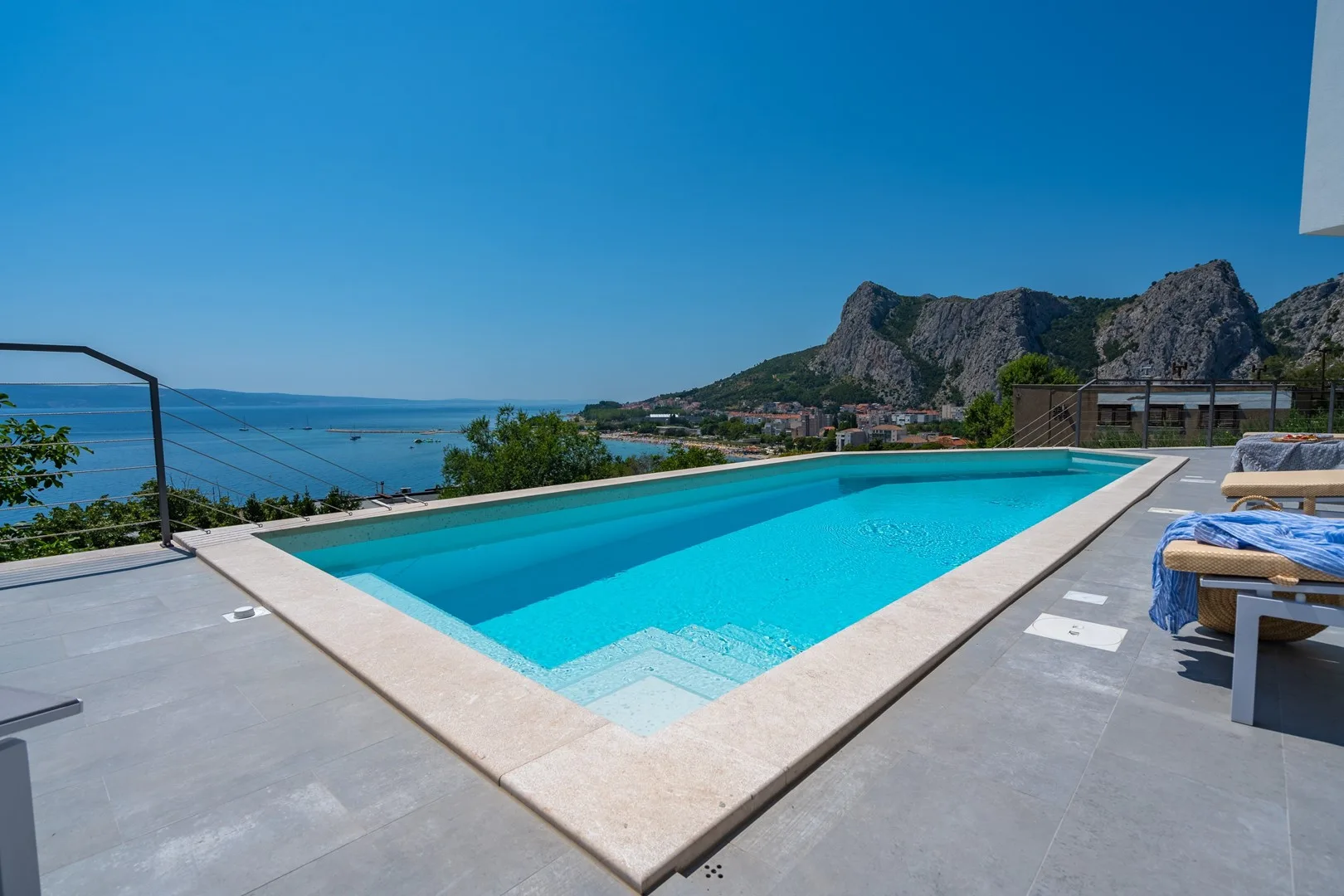An outdoor swimming pool overlooks the sea and rugged mountains.