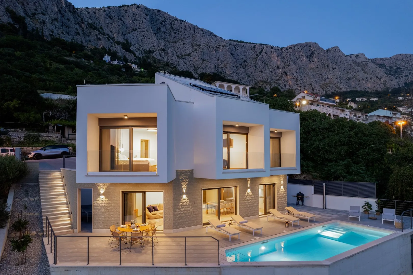 A modern house with a swimming pool sits below a rocky mountain at dusk.