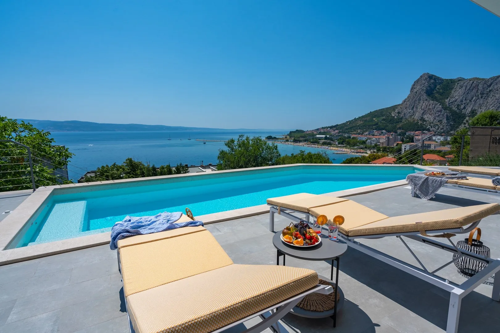 A swimming pool and sun loungers overlook the blue water and distant mountains.
