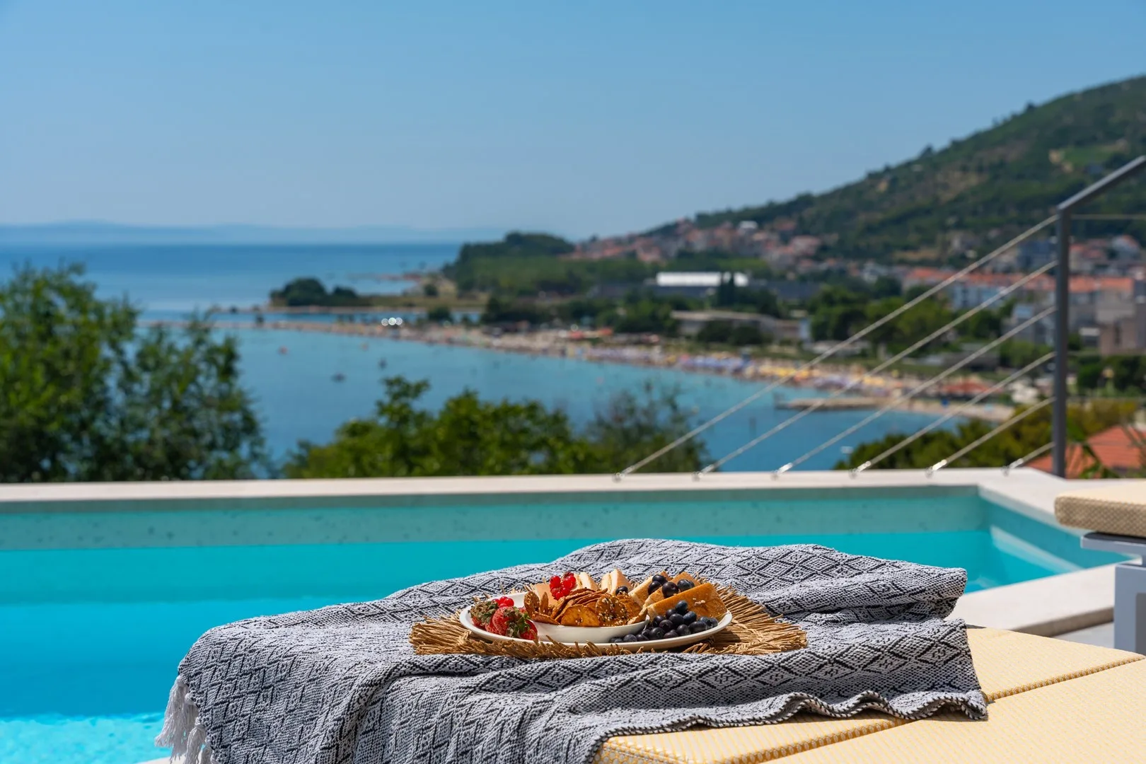 A poolside table holds a meal overlooking the sea and a coastal hillside.