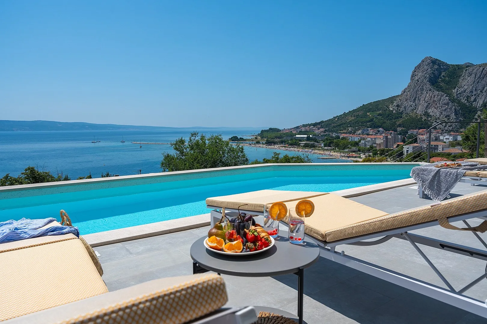 A poolside seating area overlooks the sea and a coastal town.