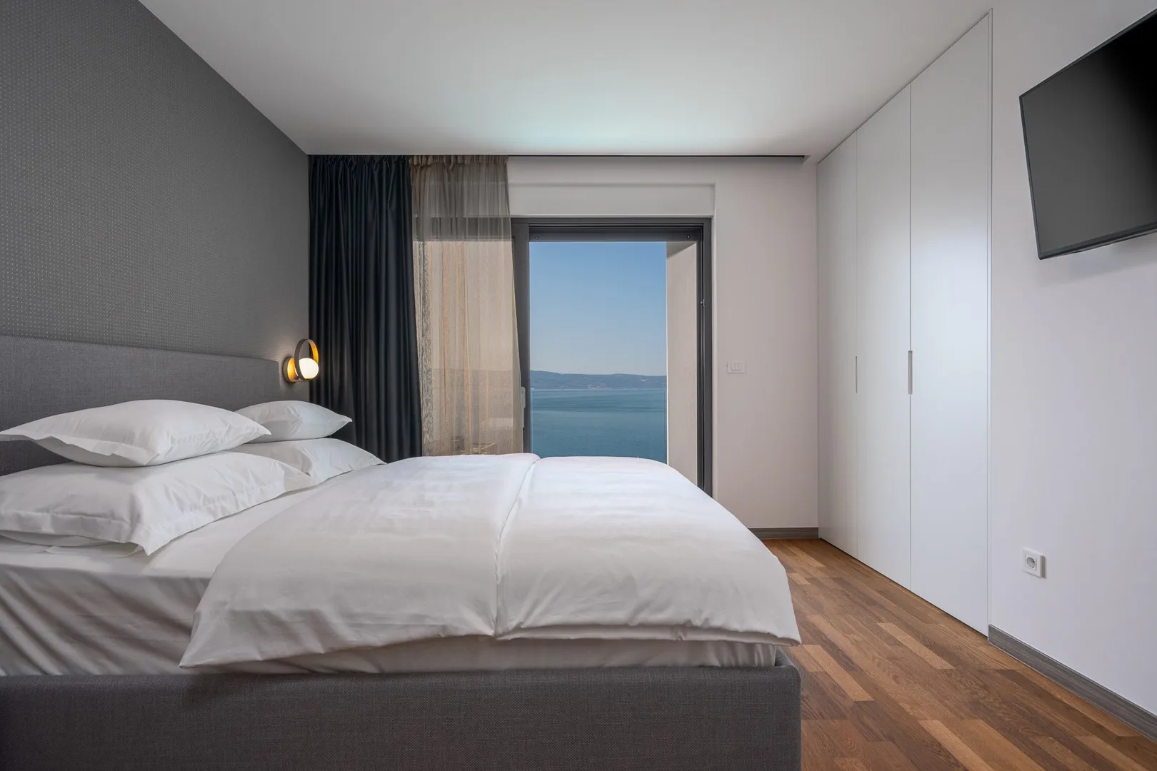 A bedroom with a neatly made bed, floor-to-ceiling curtains, and a view of the water.
