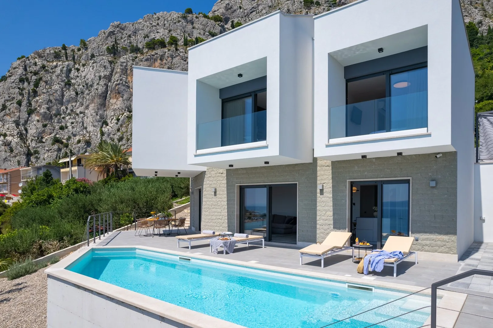 A modern white villa stands beside a swimming pool, with rocky hills behind it.
