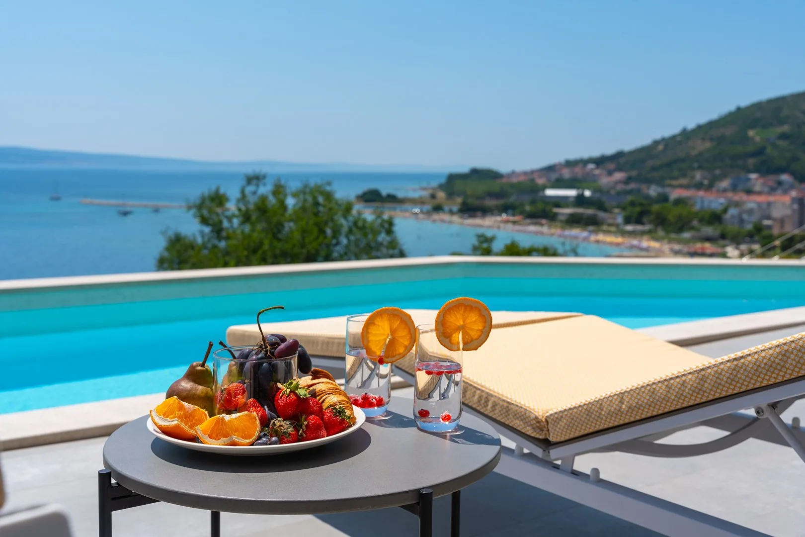 A poolside table holds a fruit platter and drinks beside a sun lounger.