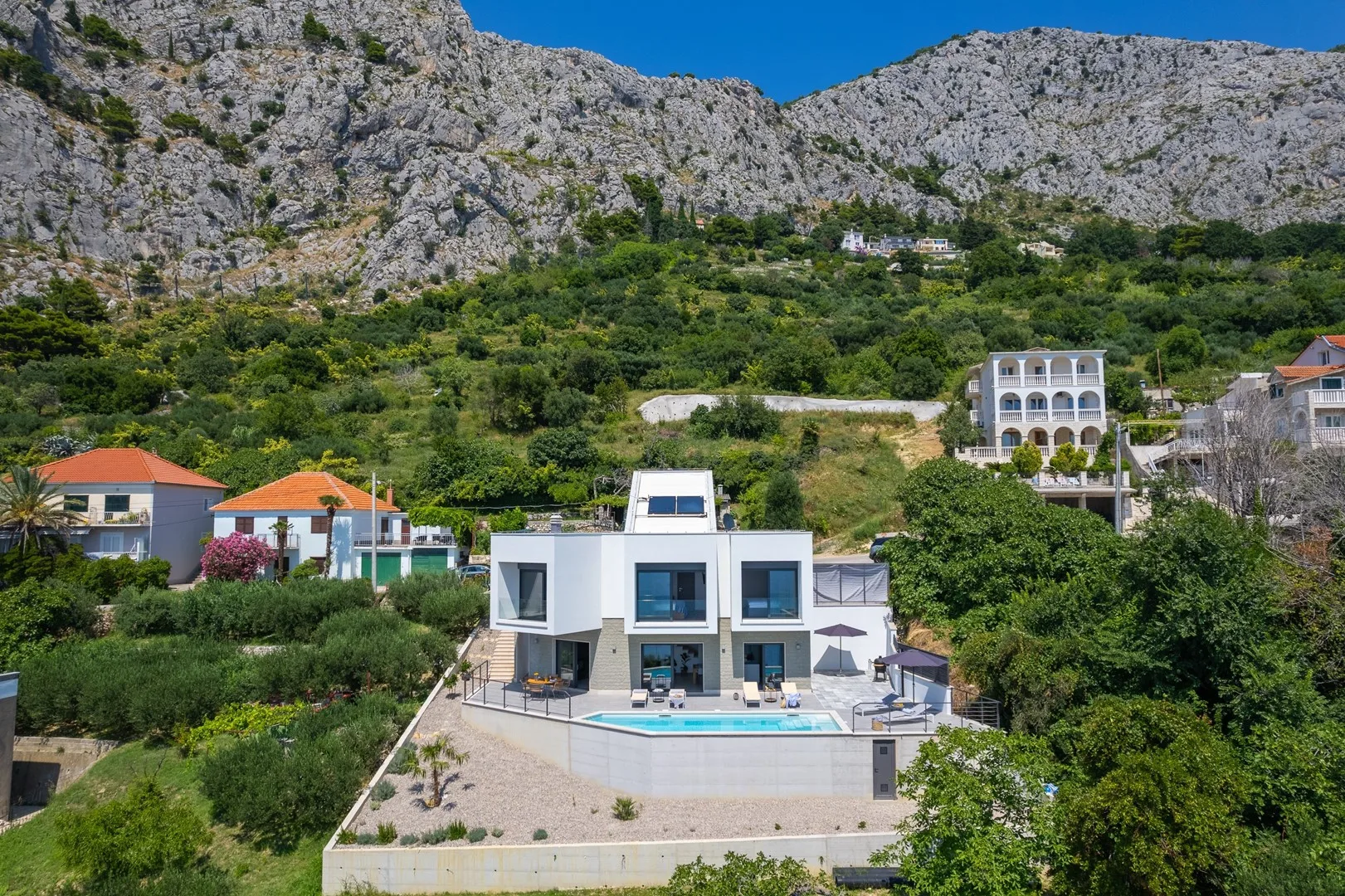 A modern house with a swimming pool sits among greenery beneath rocky mountains.