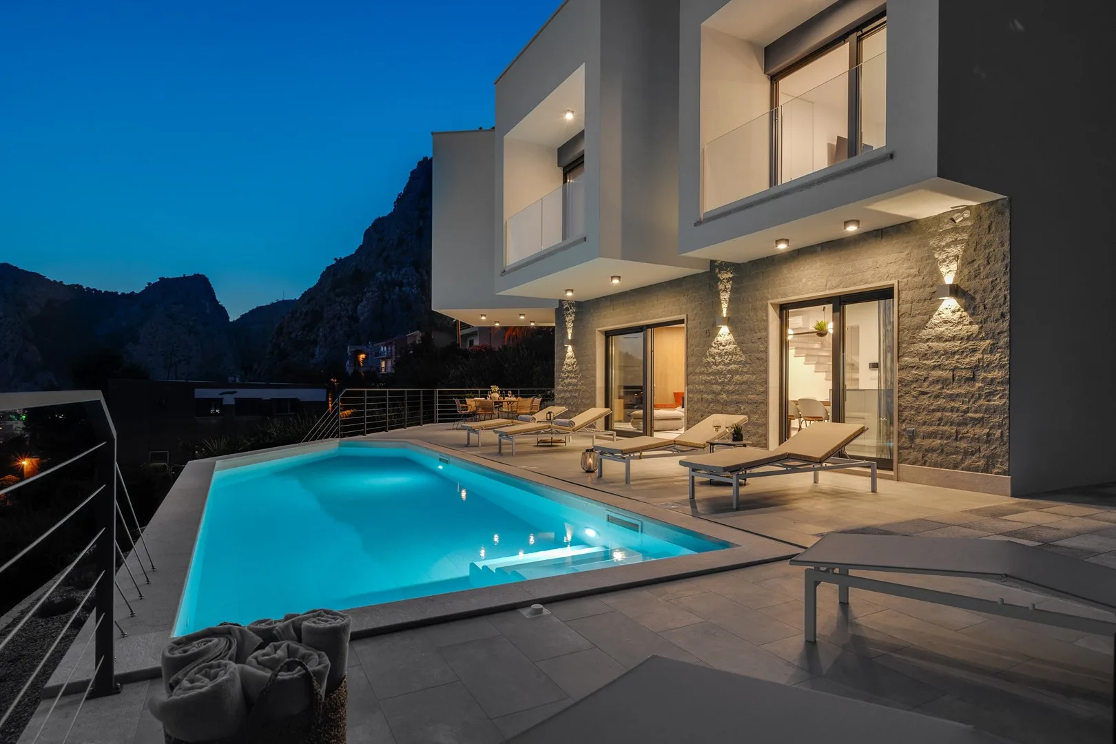 A modern house overlooks a lit swimming pool and outdoor seating at dusk.