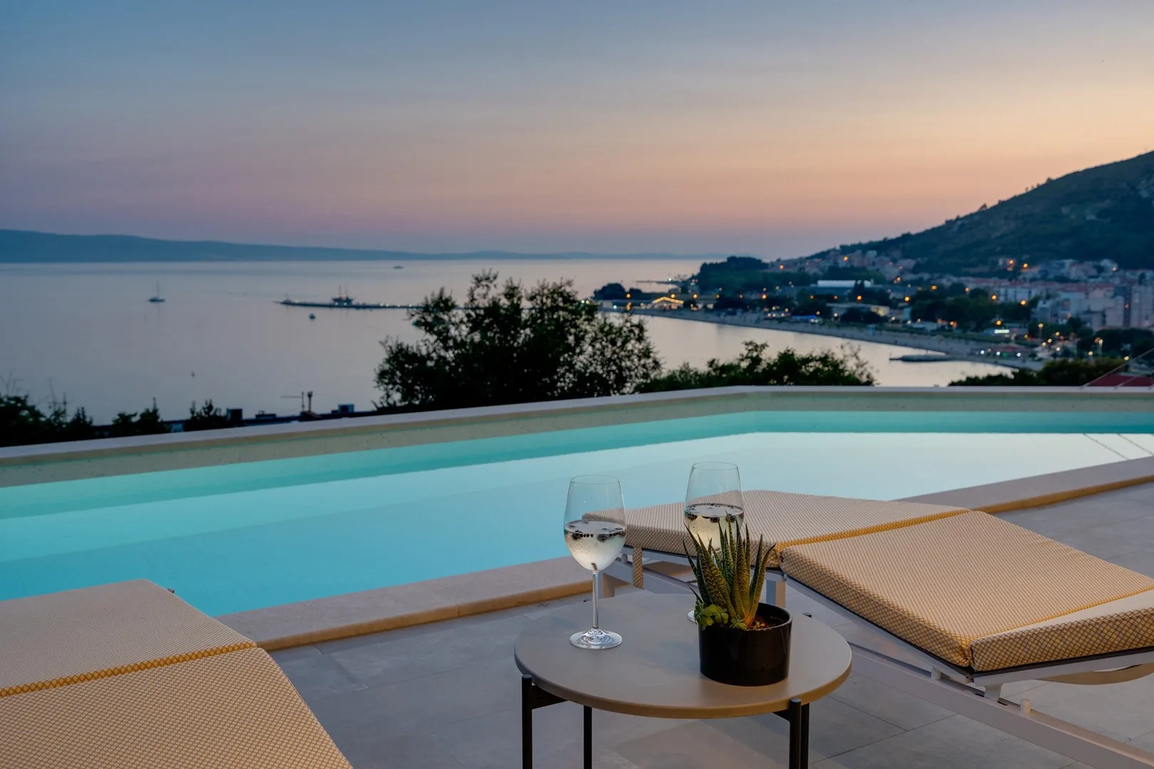 An outdoor pool overlooks a calm bay and a hillside town at dusk.