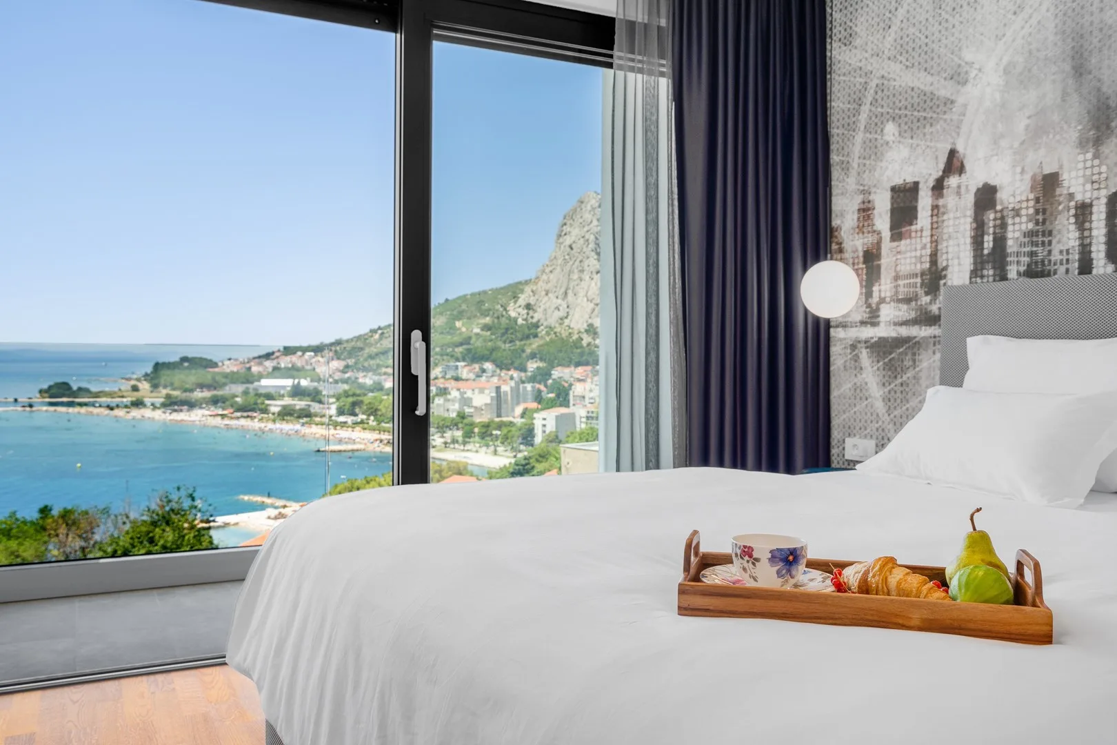 A bedroom with a white bed and a large window overlooking the coast.