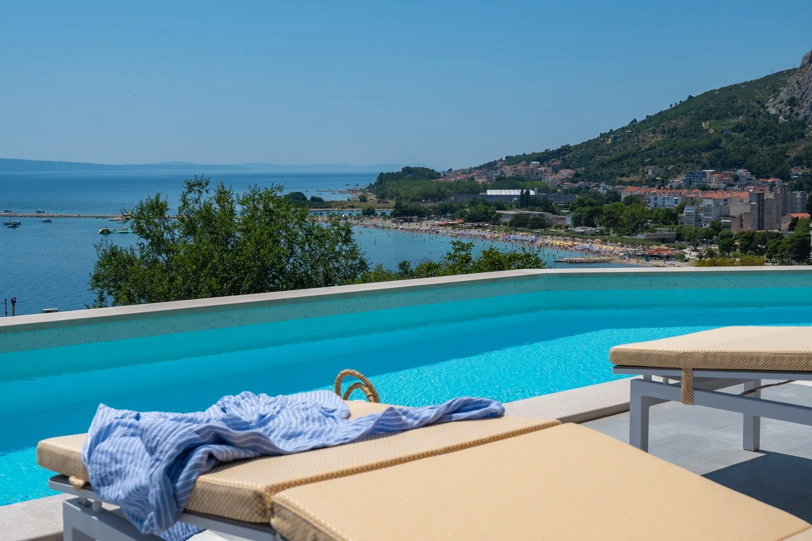 An outdoor swimming pool overlooks the sea, with cushioned loungers beside it.
