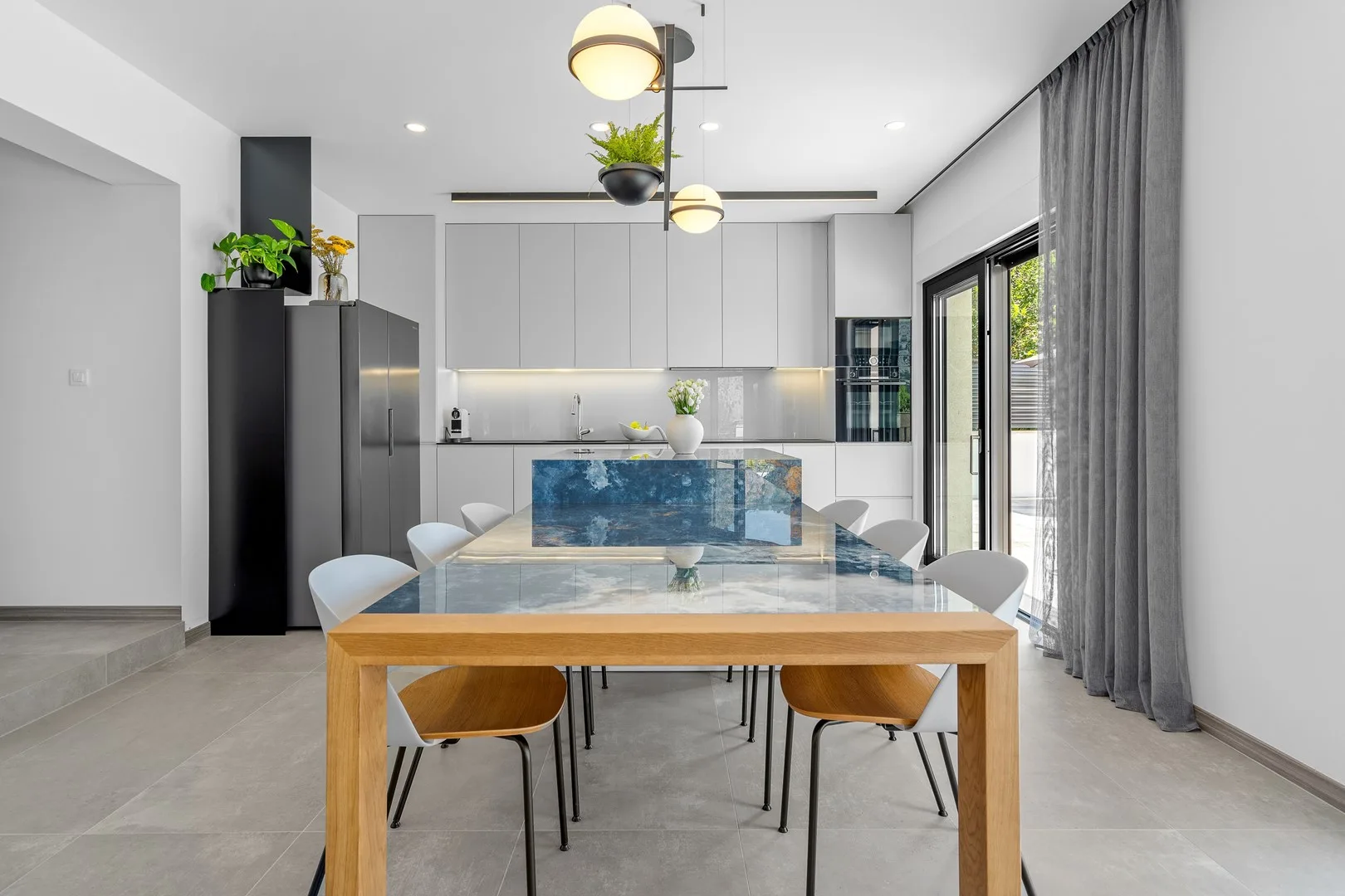 A bright modern dining area with a long table and kitchen behind it.