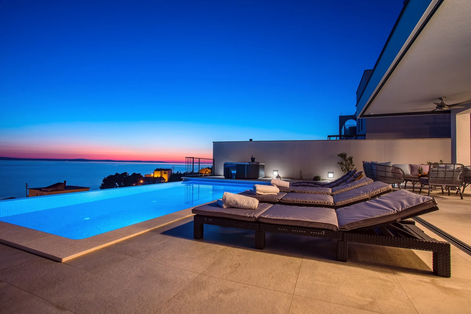 A poolside terrace overlooks the sea at sunset, with lounge seating beside the water.