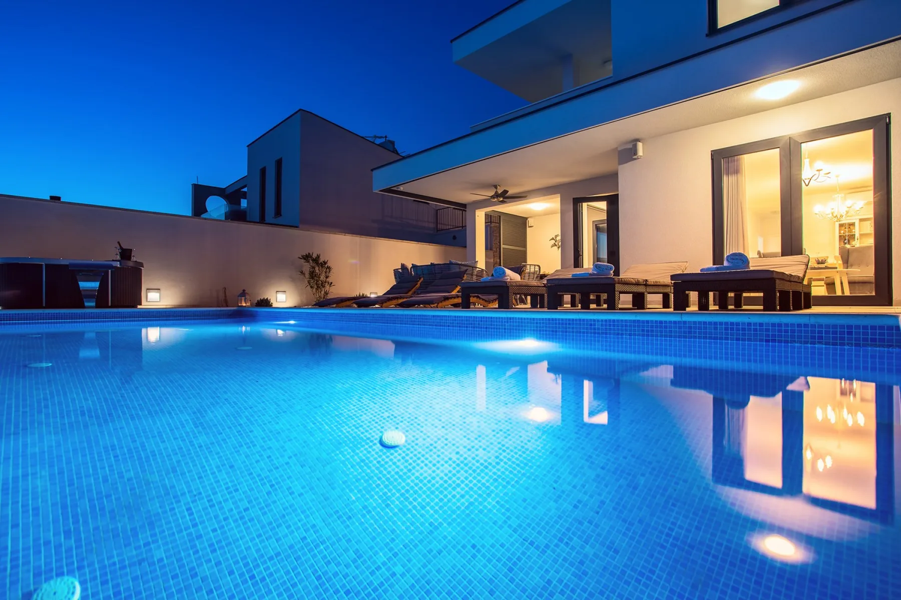 An illuminated swimming pool beside a modern house at dusk.