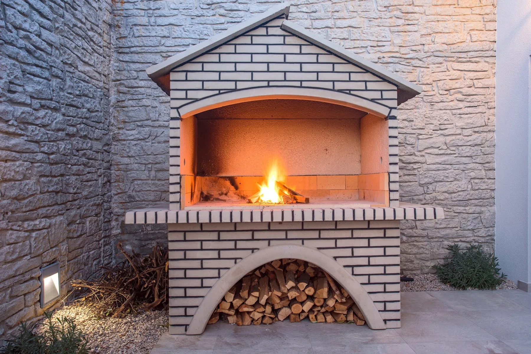 A brick outdoor fireplace burns beside a stack of firewood.