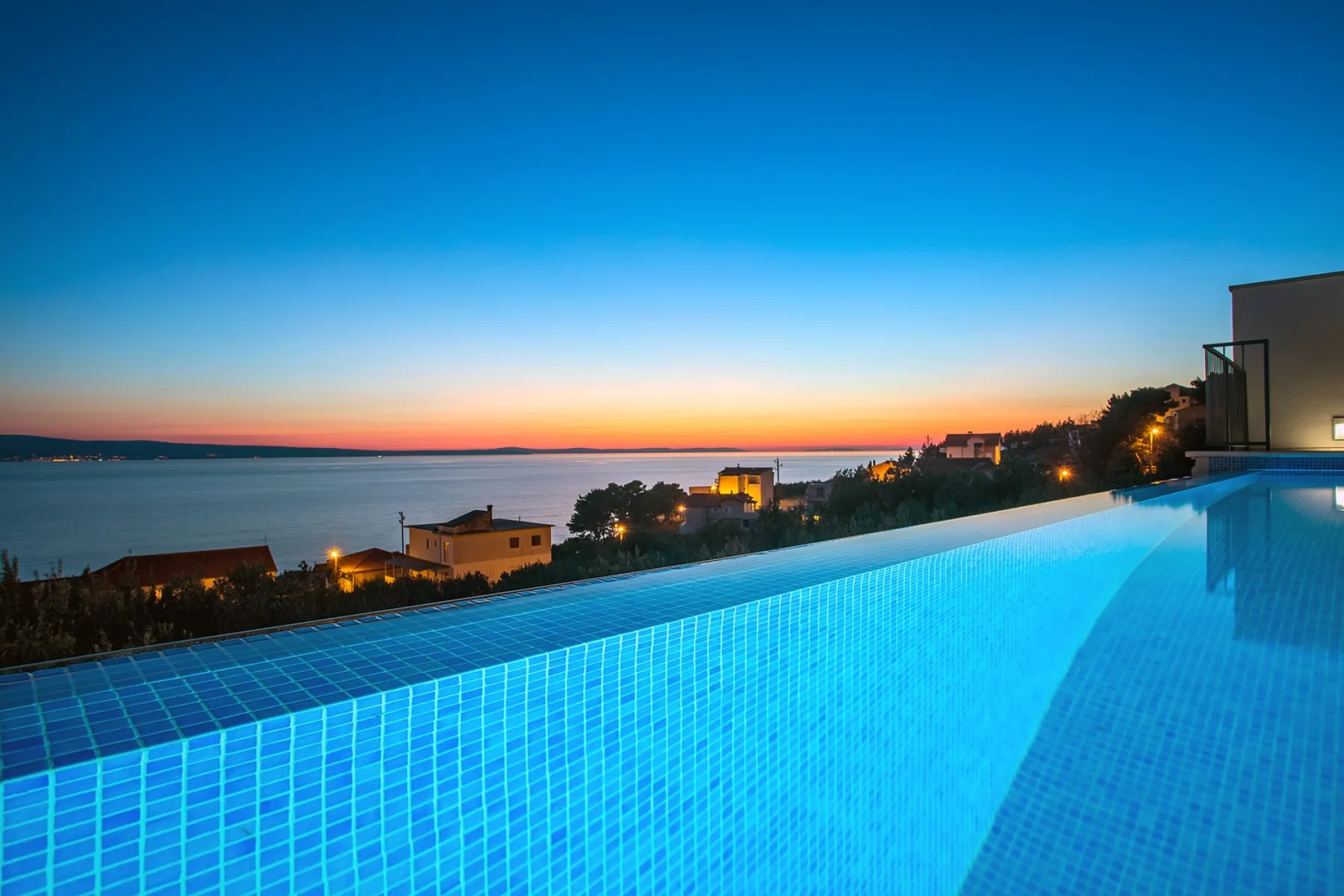 An illuminated swimming pool overlooks the water at sunset.