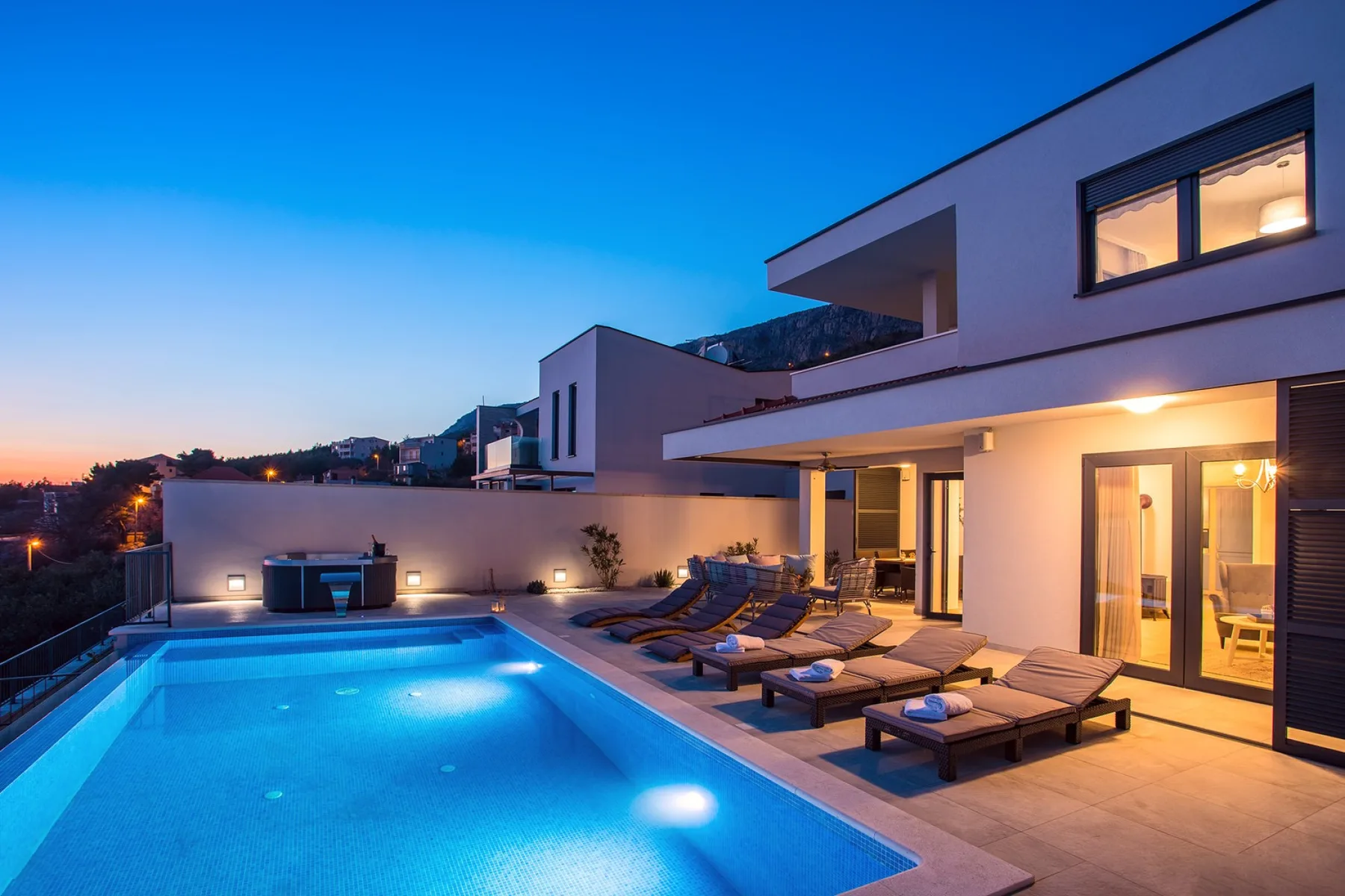 A swimming pool and lounge chairs sit beside a modern house at dusk.