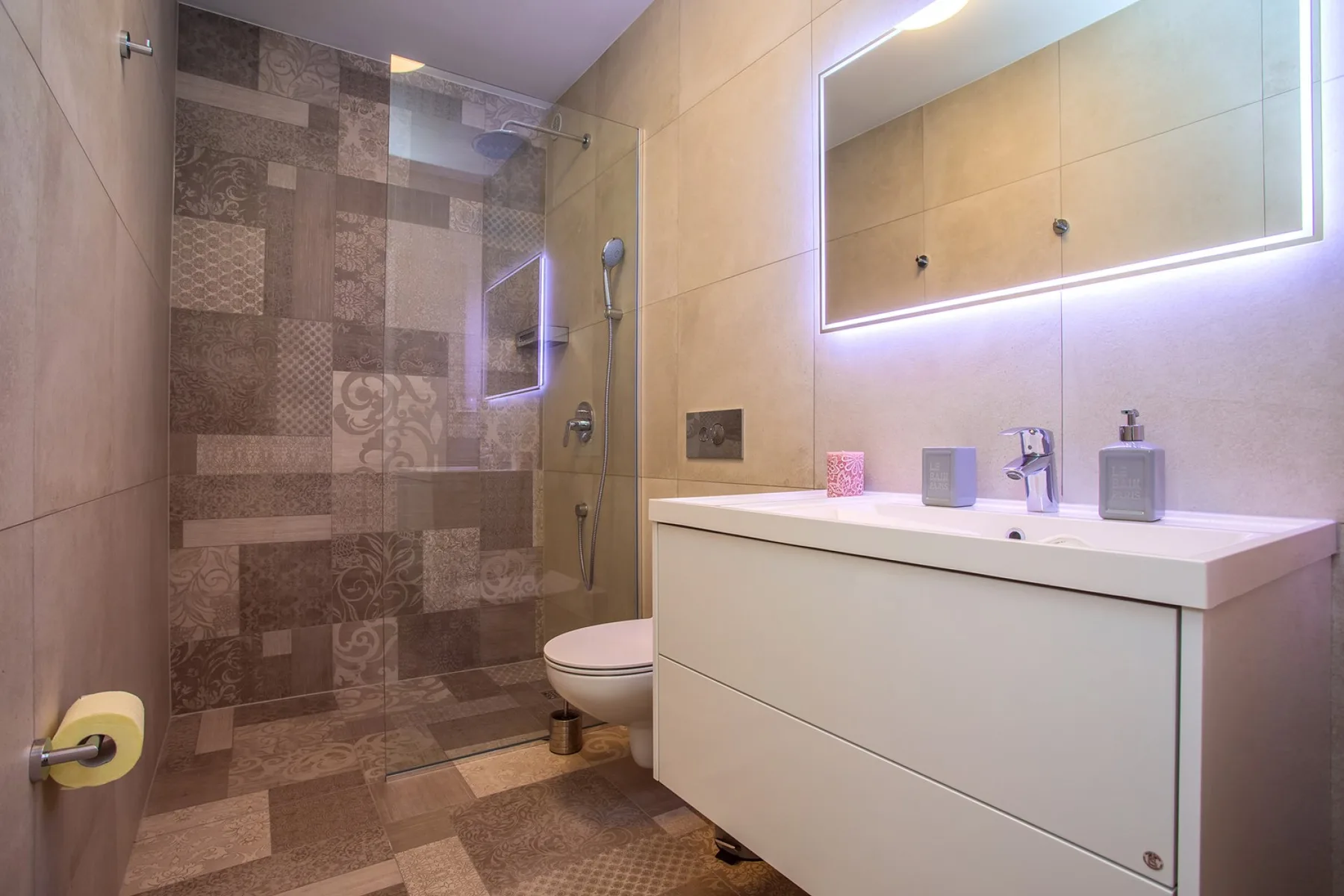 A tiled bathroom with a glass shower, toilet, and vanity beneath a lit mirror.