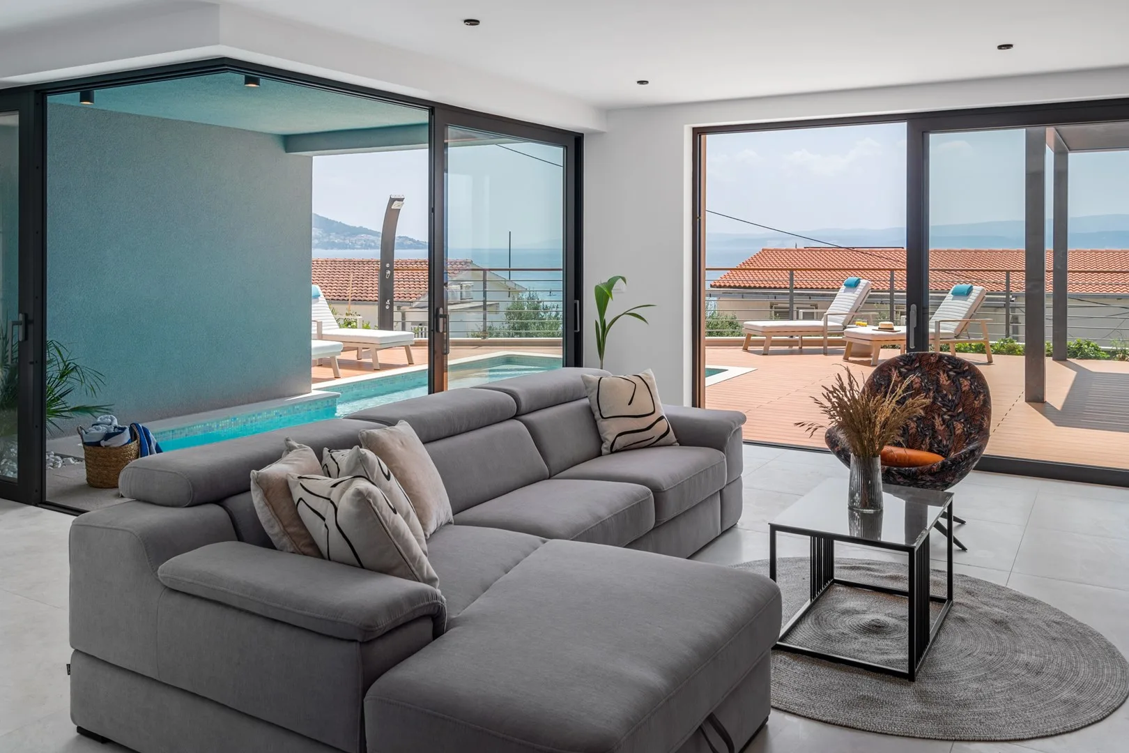 A living room with gray seating opens onto a patio overlooking the sea.