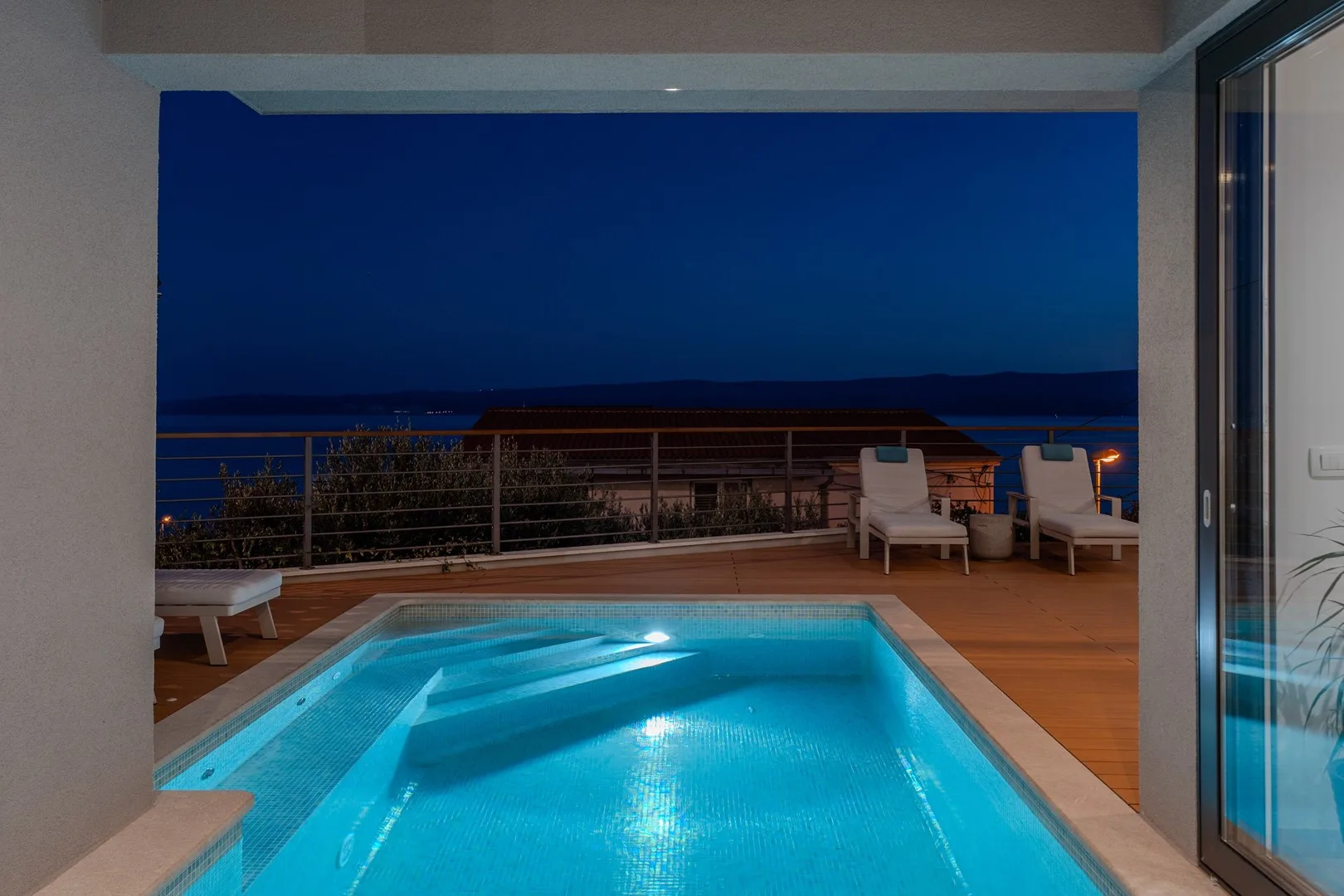 A lit swimming pool sits beside a wooden deck overlooking the water at dusk.