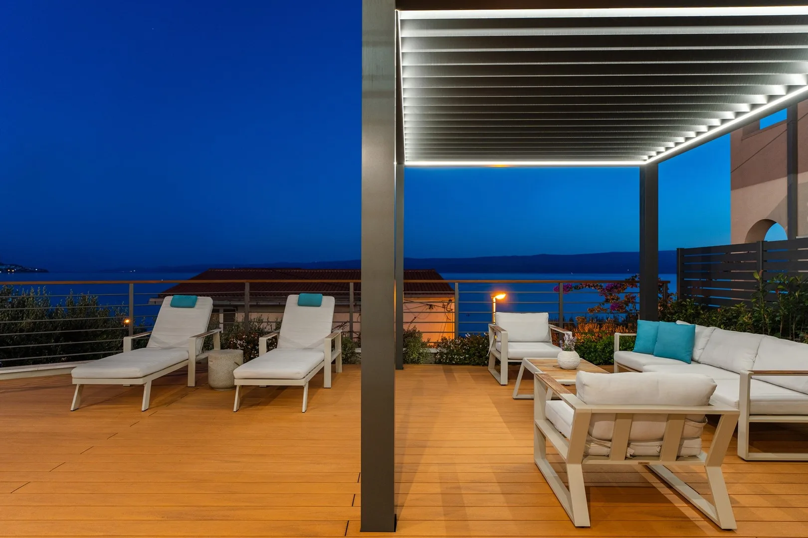 An outdoor terrace with lounge seating beneath a lit pergola overlooking the sea.