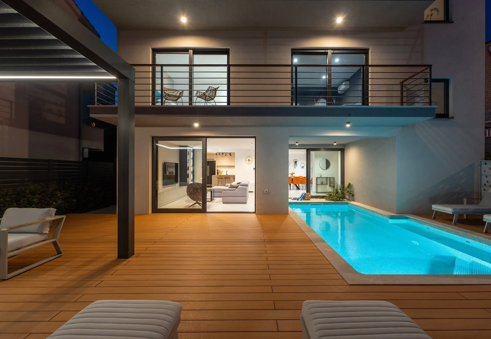 A modern house opens onto a wooden deck beside a brightly lit swimming pool.