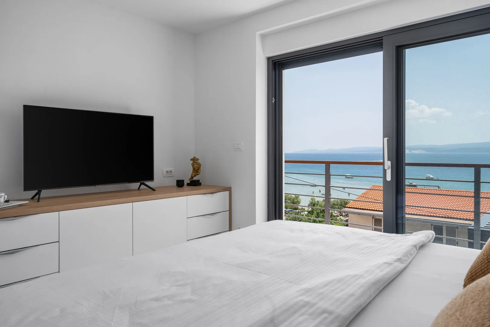 A bright bedroom with a bed, television, and large windows overlooking the sea.