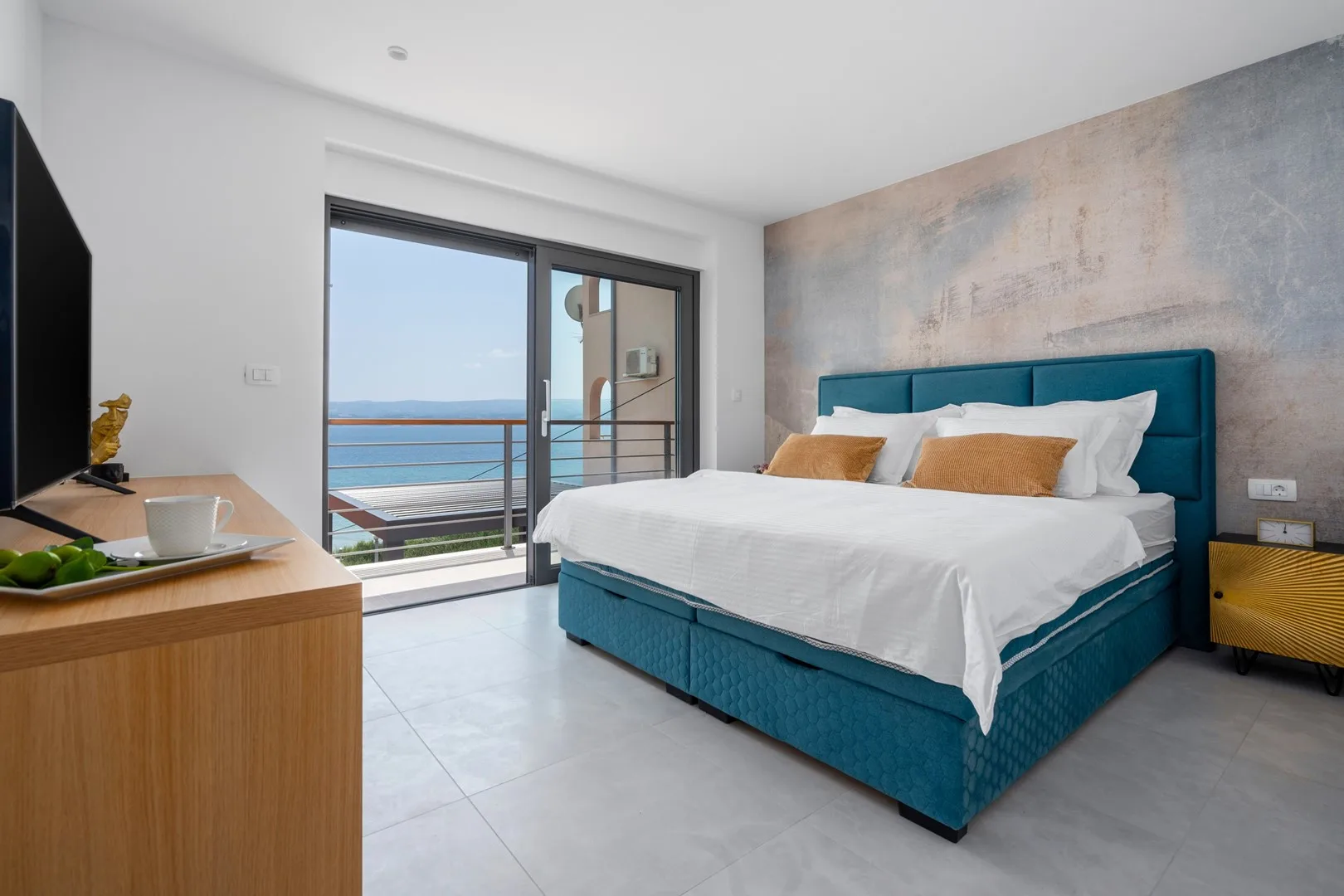 A bedroom with a teal bed beside sliding glass doors overlooking the sea.