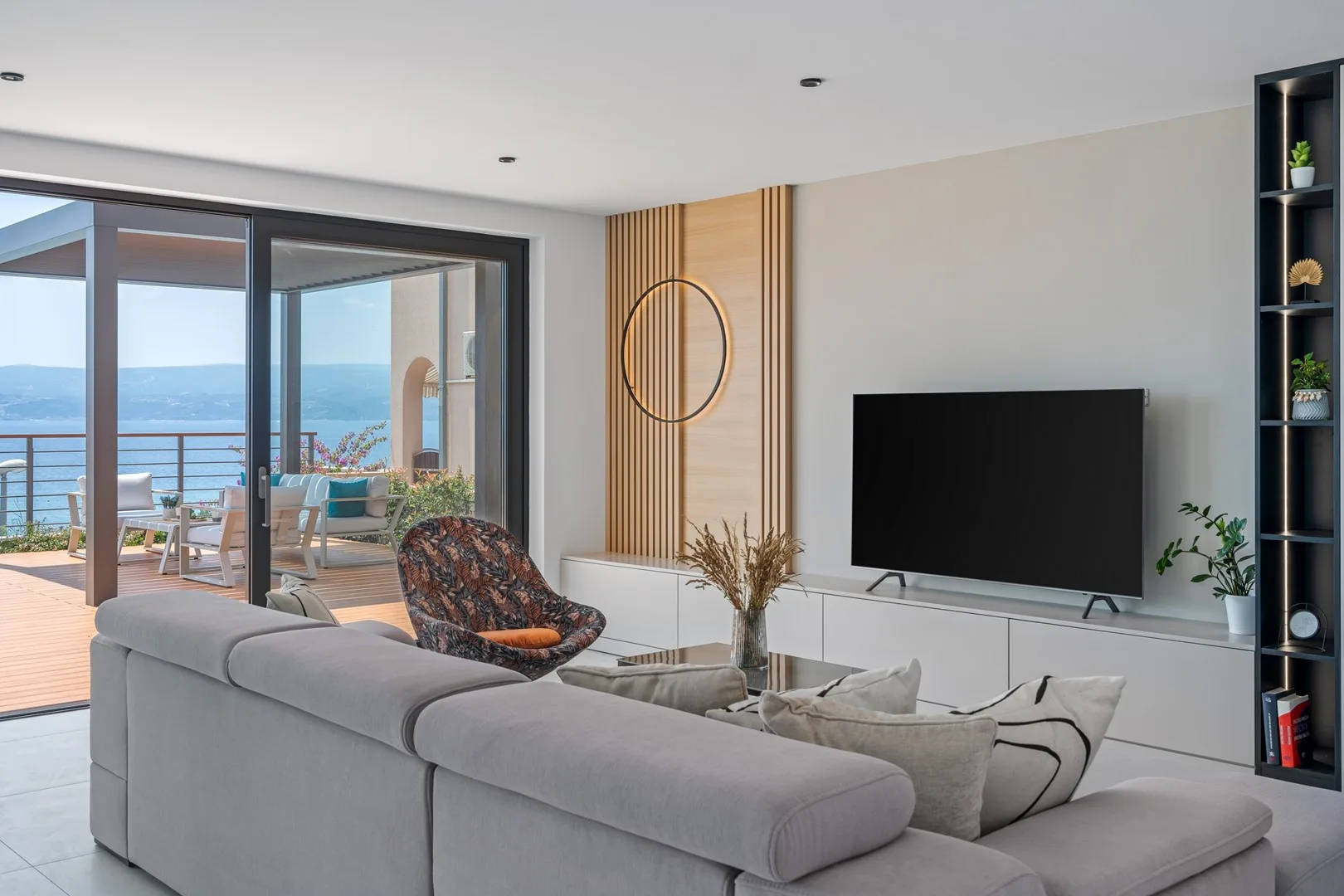 A bright living room with seating, a television, and large windows overlooking the sea.