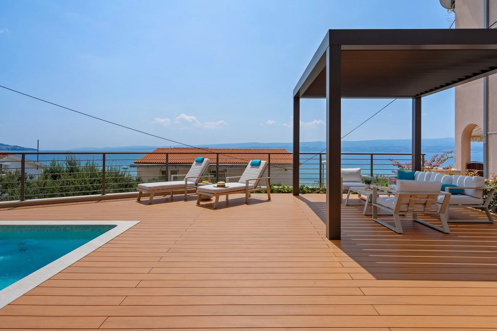 A poolside deck overlooks the sea, with lounge seating beneath a pergola.