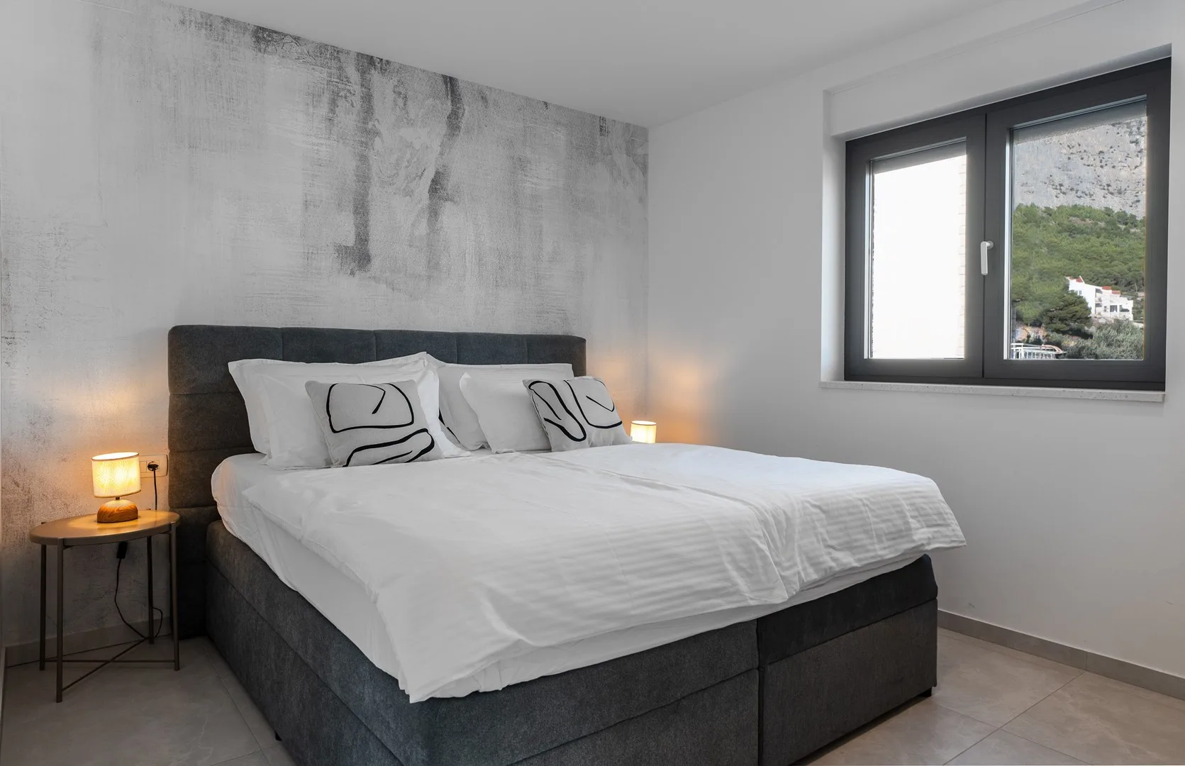 A bedroom with a large bed, bedside tables, and a window overlooking greenery.