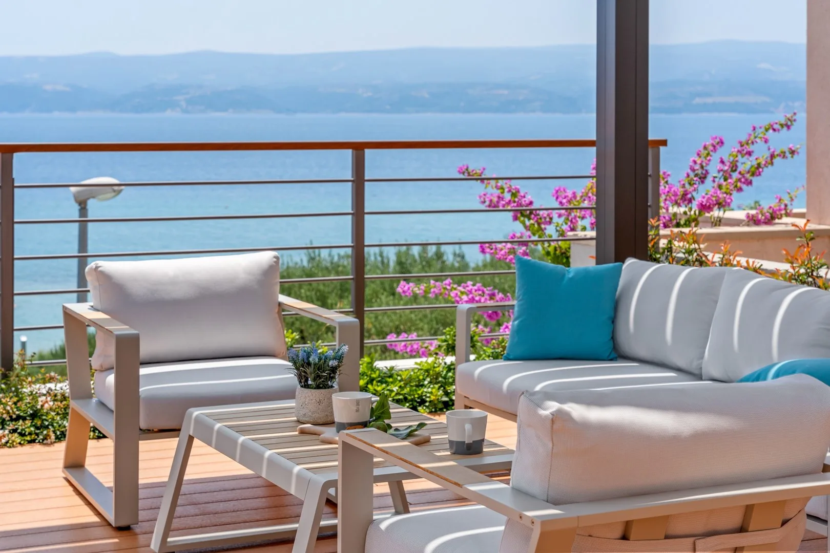 An outdoor seating area overlooks the water, with flowering plants beside the railing.