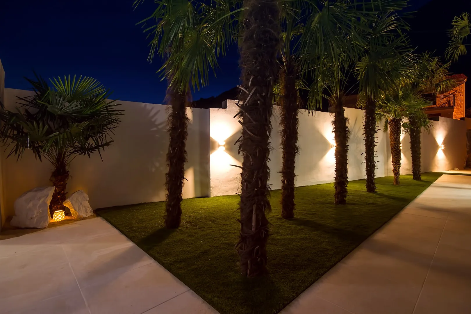 Un patio exterior ajardinado con palmeras, césped y muros iluminados por la noche.