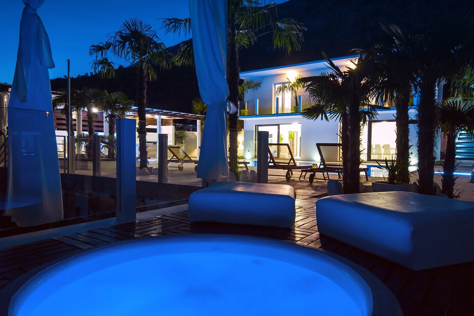Una zona de piscina exterior iluminada, con asientos y palmeras por la noche.