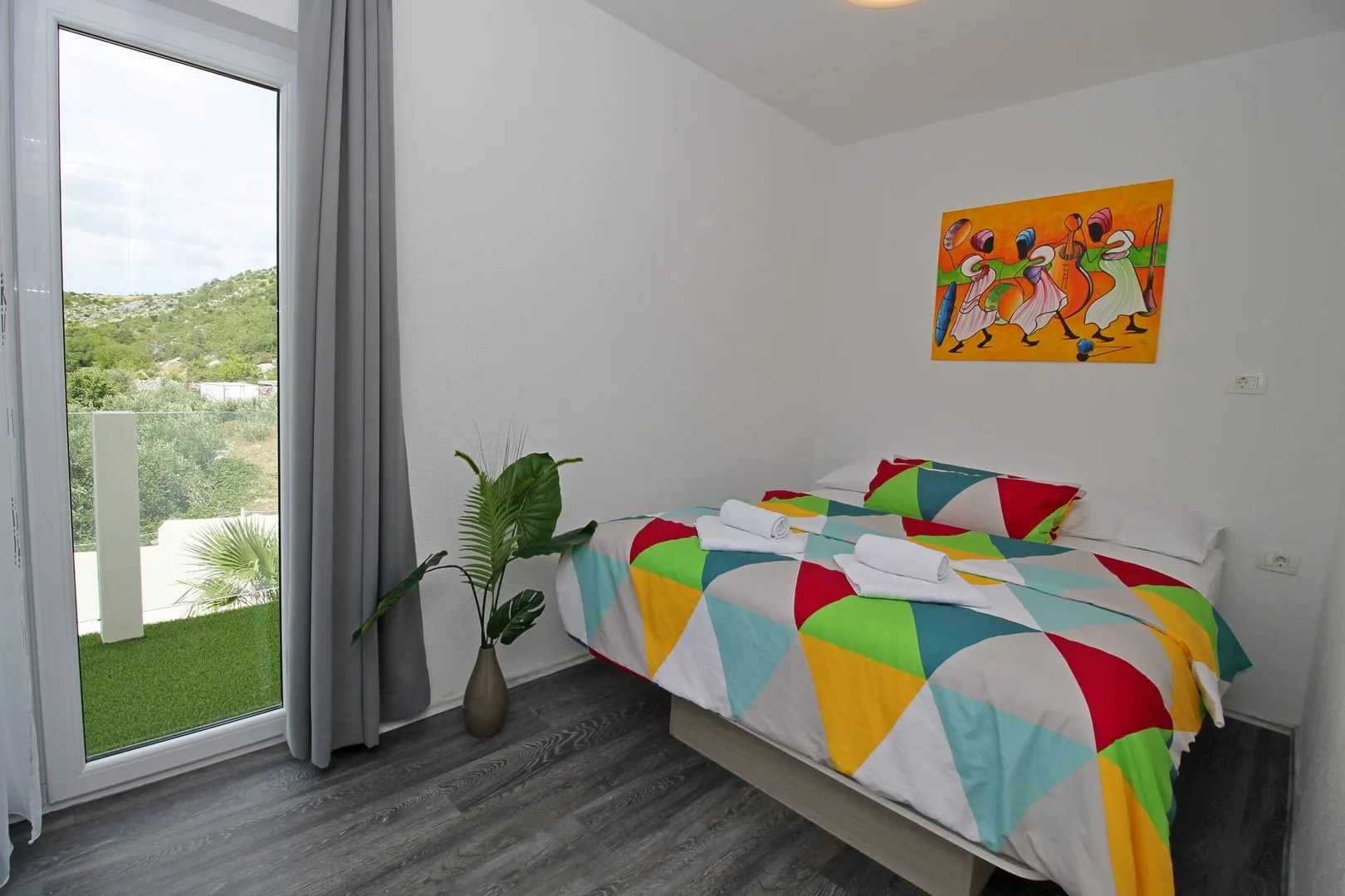 Un dormitorio con ropa de cama geométrica y colorida, una planta y una puerta de vidrio hacia el exterior.