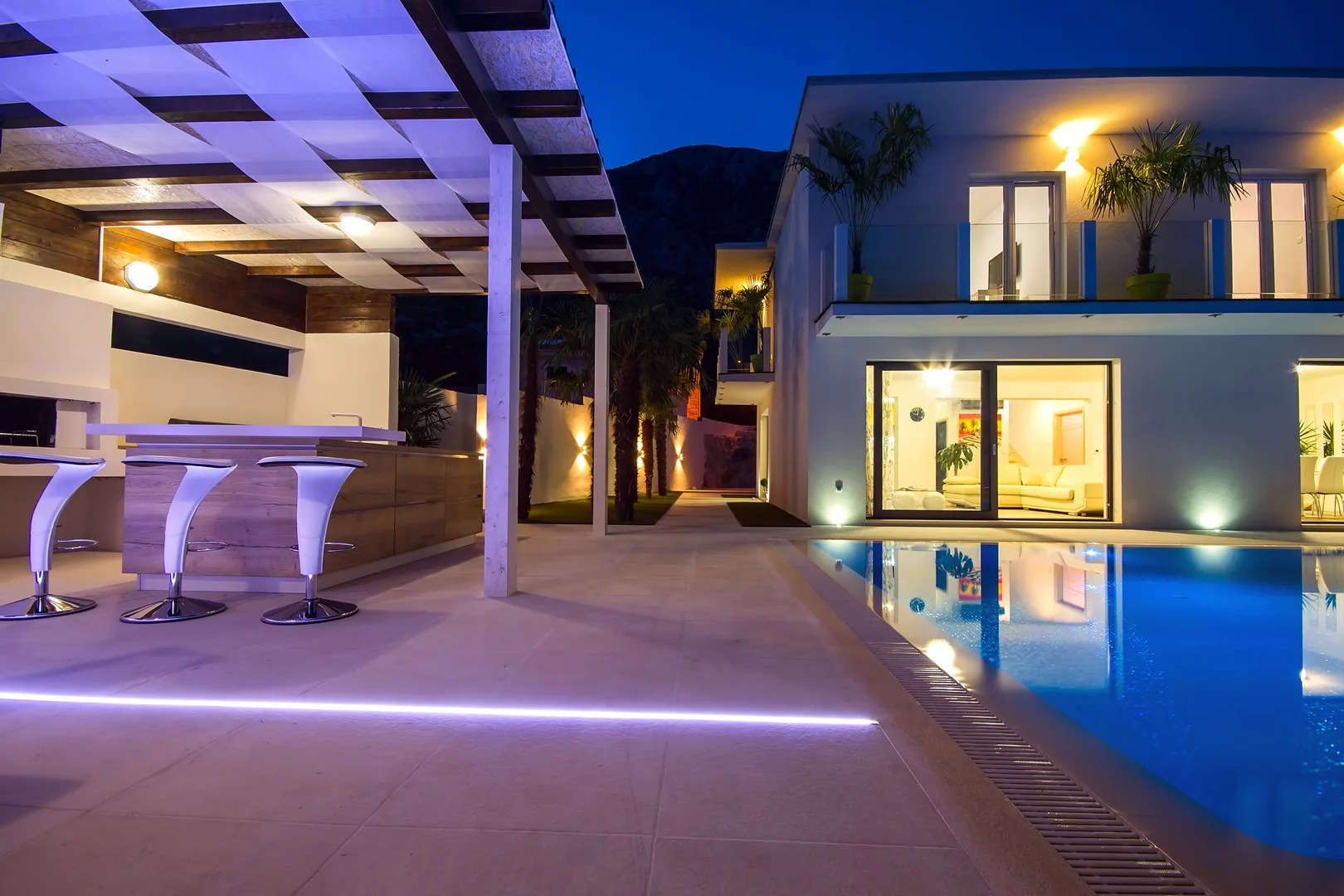Una casa moderna y una piscina exterior están iluminadas al anochecer junto a un patio cubierto.