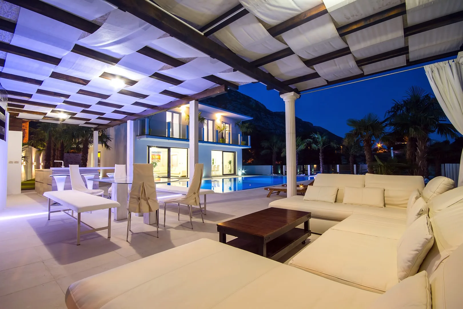 Una zona exterior de estar y comedor da a una piscina iluminada bajo un patio cubierto.