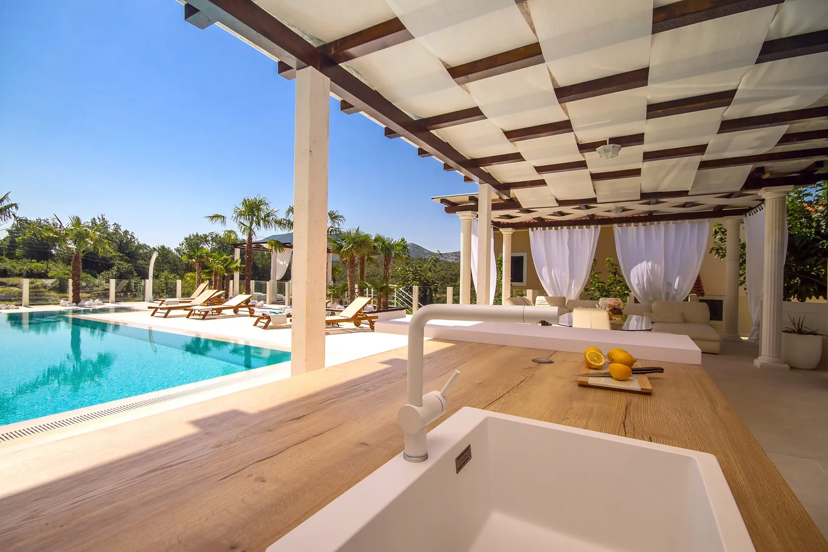 Una zona de comedor junto a la piscina cuenta con una encimera de madera bajo una pérgola sombreada.
