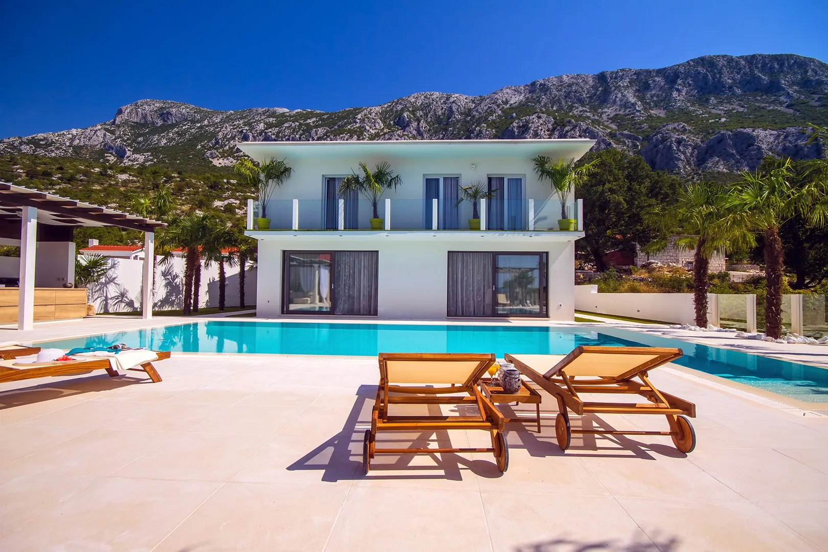 Una casa moderna da a una piscina y tumbonas, con montañas al fondo.
