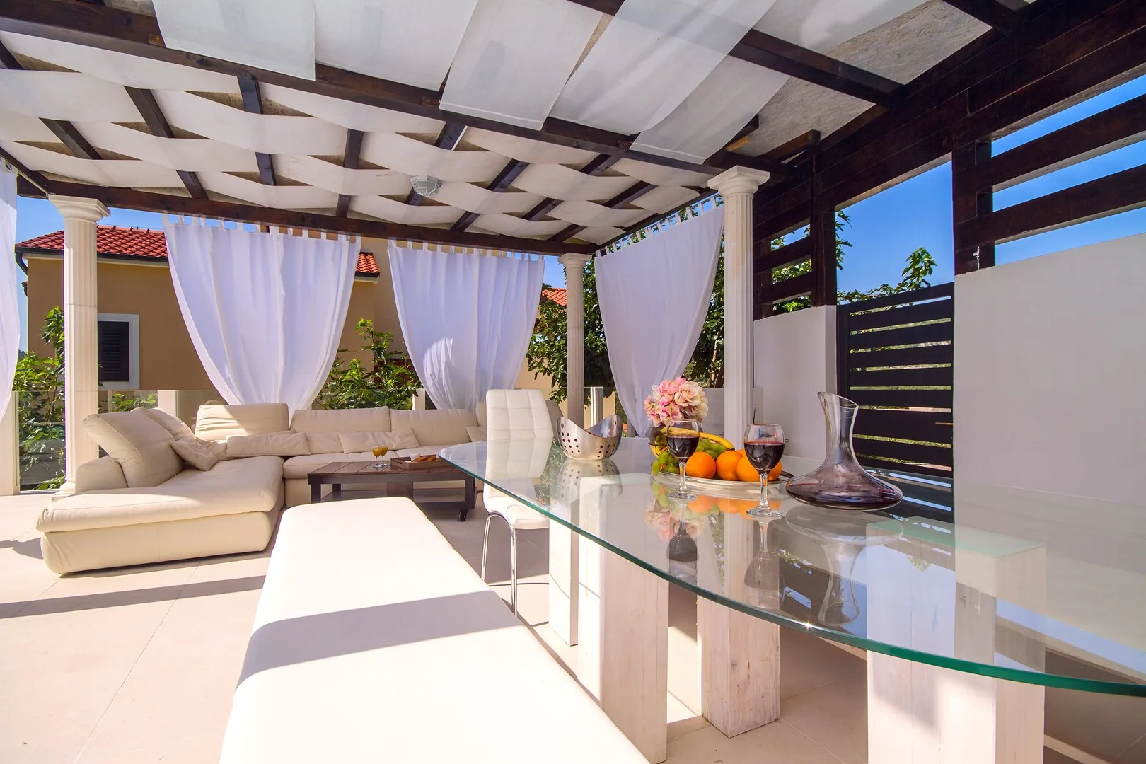 Un patio cubierto y luminoso tiene asientos, cortinas translúcidas y una mesa de comedor de vidrio.