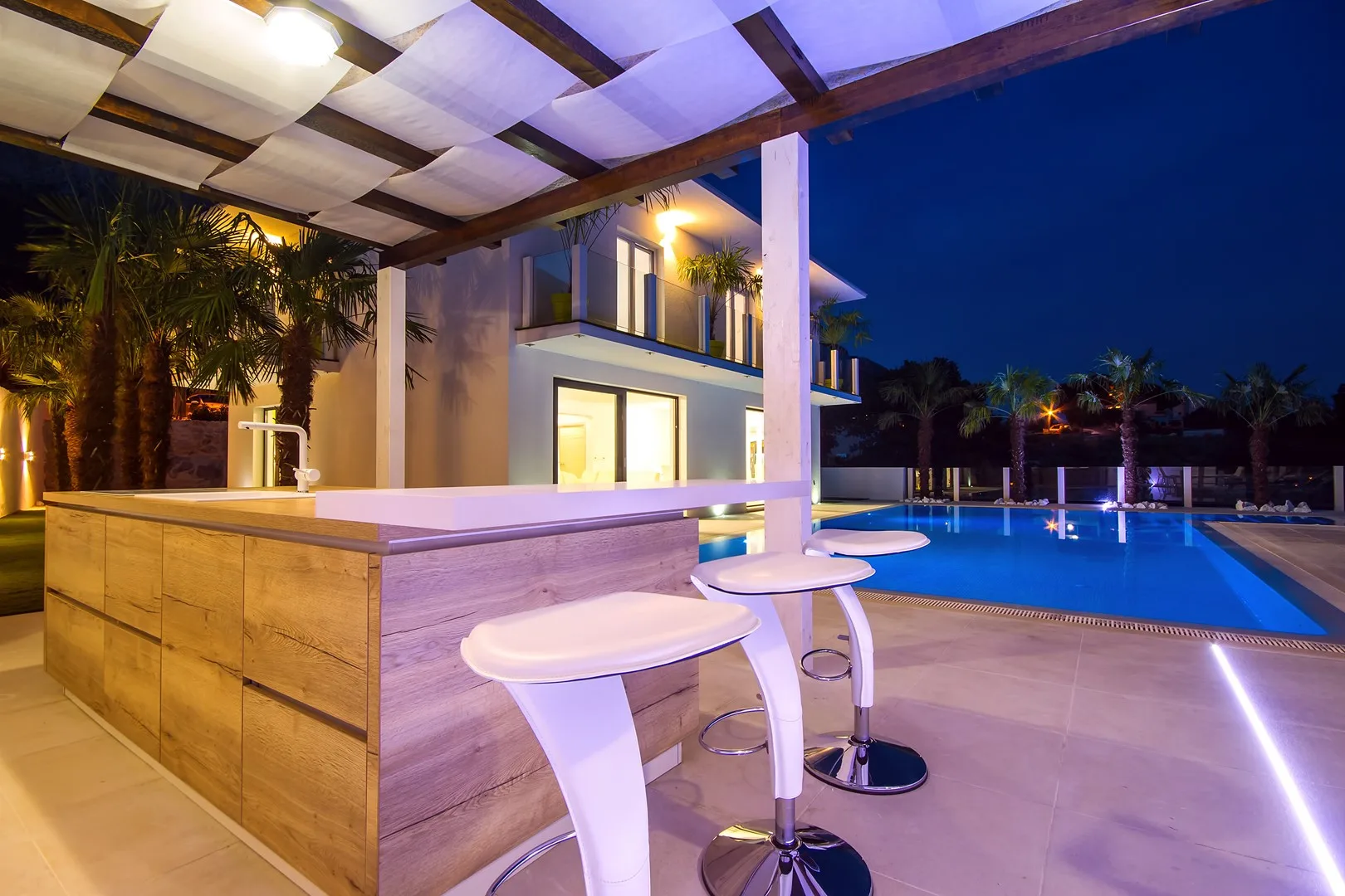 Una barra exterior con asientos da a una piscina junto a un edificio moderno por la noche.