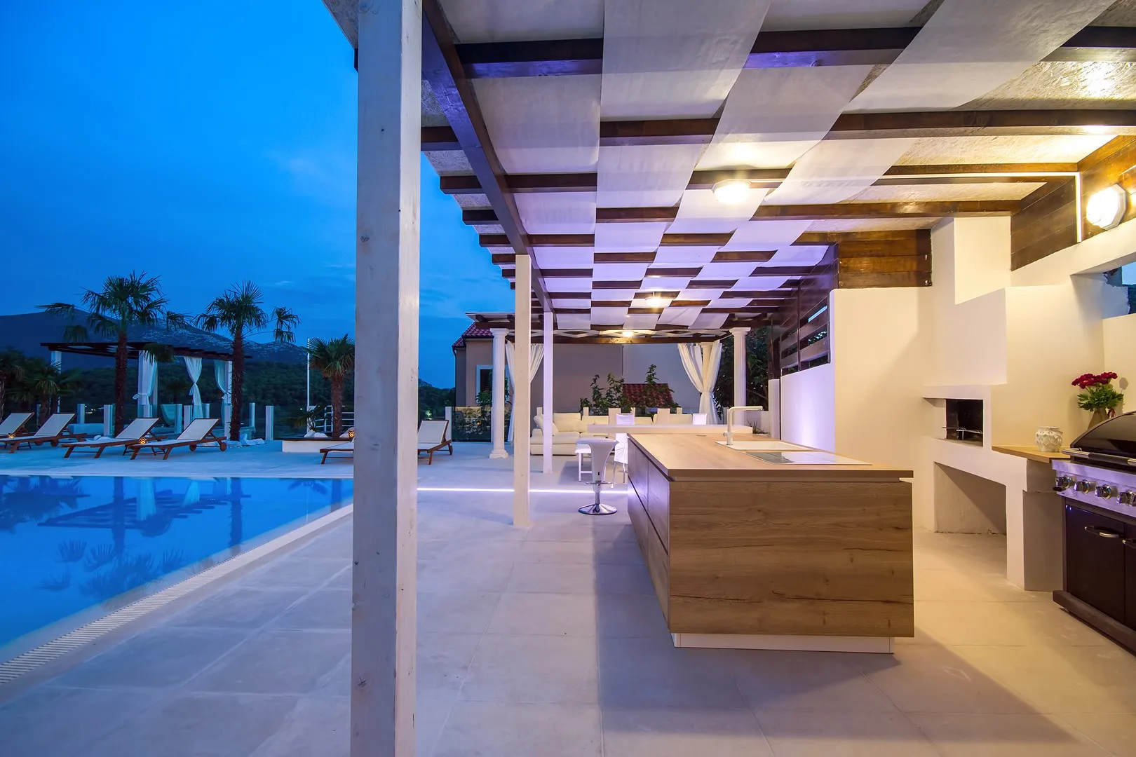 Una cocina y zona de comedor al aire libre da a una piscina al anochecer.