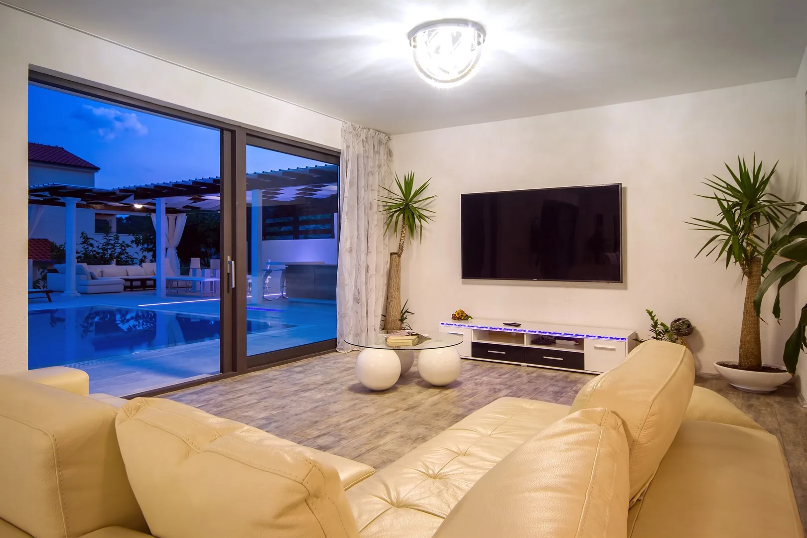 Una sala de estar con asientos frente a un televisor en la pared y grandes ventanas con vista a una piscina.